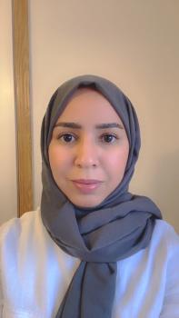 Asma Albeladi wearing a white shirt, standing in front of a tan wall.