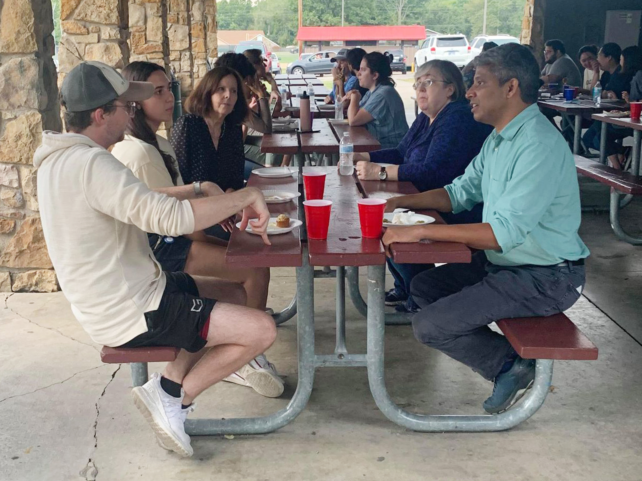 Students and faculty sit together at picnic tables inside outside shelter